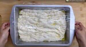 Focaccia dough rising in an oil-coated baking pan, covered with plastic wrap, on a wooden surface.